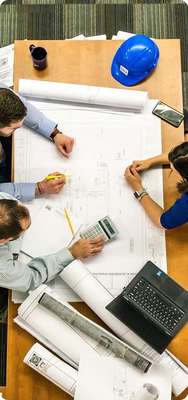 Sales engineers and team members collaborating around a table, reviewing engineering blueprints, with a hard hat, laptop, and calculator visible
